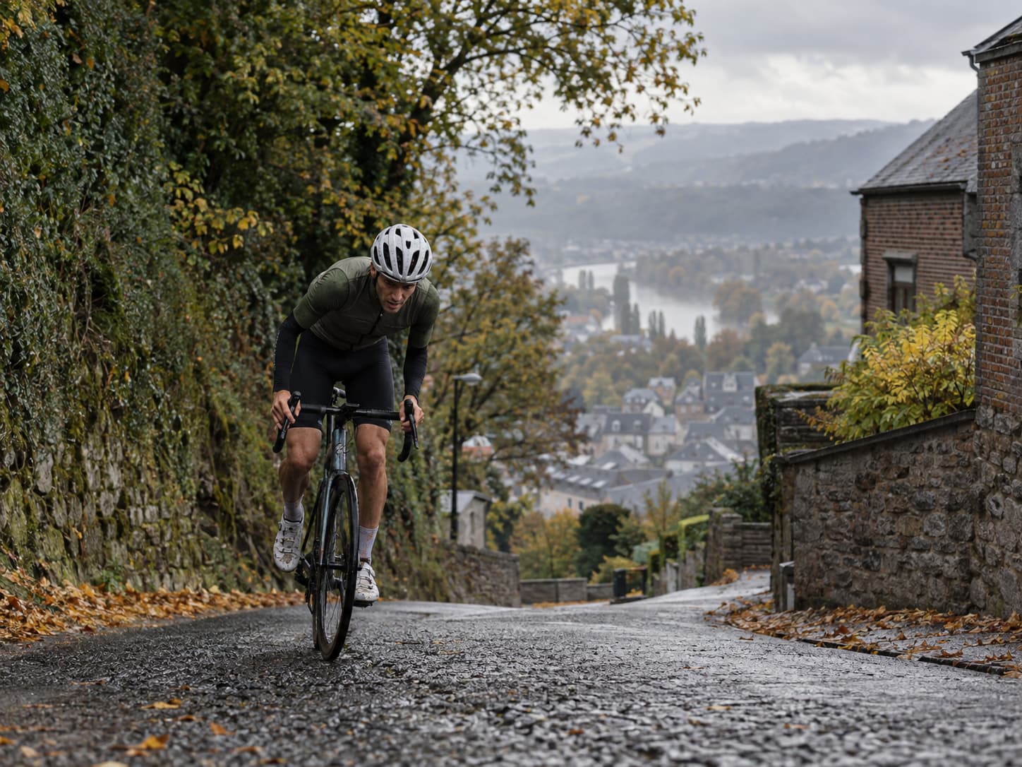 Cycliste en effort dans une forte montée asphaltée, au-dessus d'un village et d'une vallée, dans une ambiance évoquant les classiques wallonnes