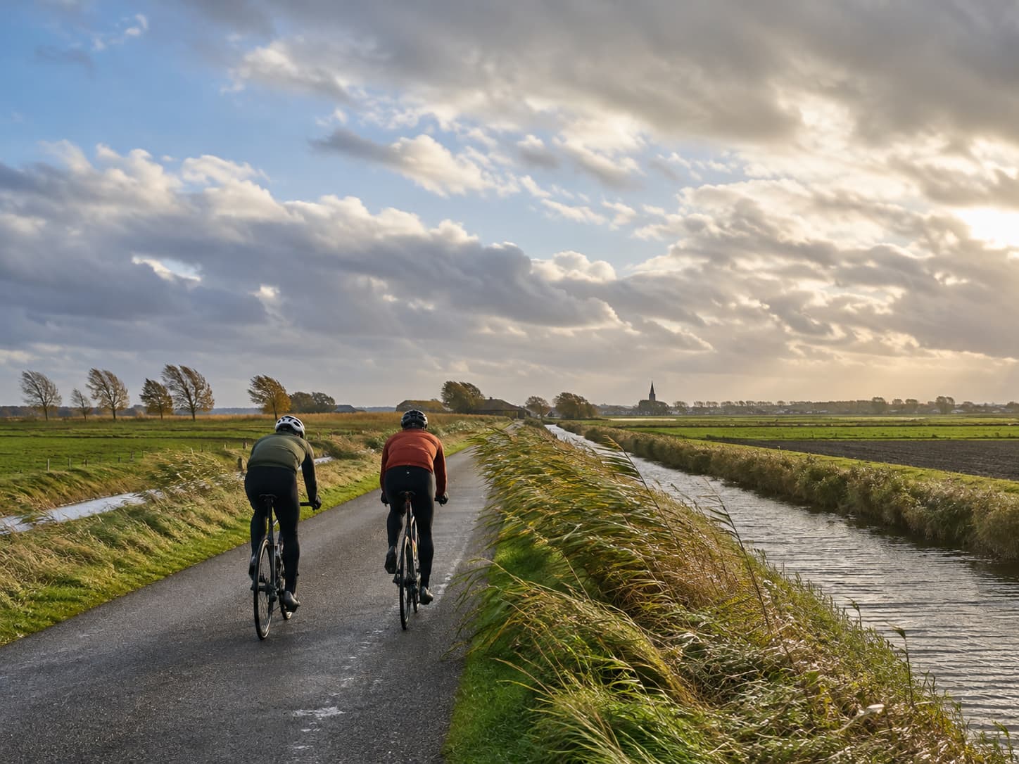 Deux cyclistes sur une route plate bordée d'un canal et de champs dans un paysage de polders flamands